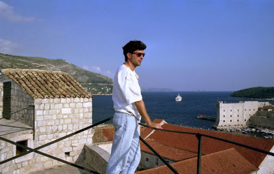 Side view of young man standing on railing against sea