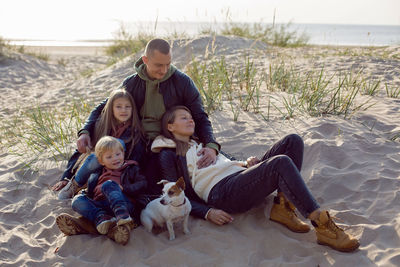 Family with a son and daughter and a dog sit on the sand in autumn in leather jackets