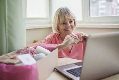 Senior woman holding heart shape using laptop at home