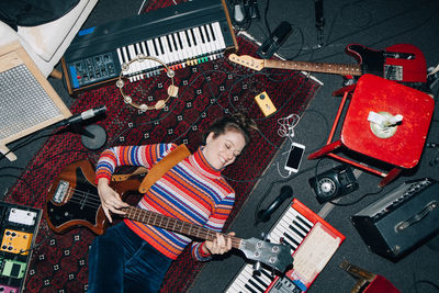 High angle view of smiling woman playing guitar while lying amidst musical instruments at home