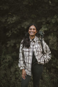 Portrait of smiling woman with hands in pockets standing in forest