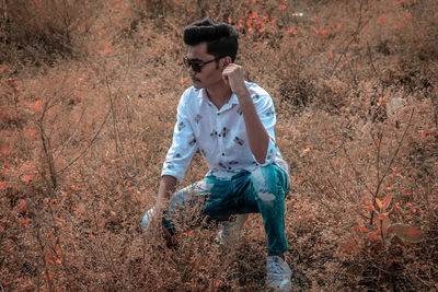 Full length of young man wearing sunglasses while crouching on land by plants