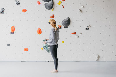 Full body side view of sportive barefoot female climber putting on safety belay near artificial climbing rock before training in gym