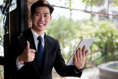 Portrait of smiling young businessman showing thumbs up while holding using digital tablet