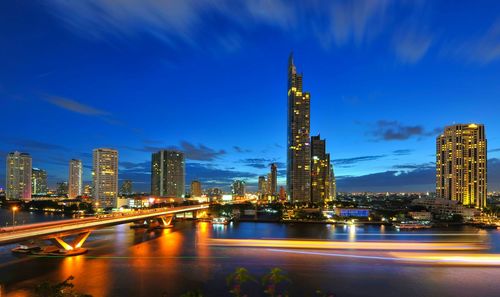 Taksin bridge over river by illuminated buildings against blue sky