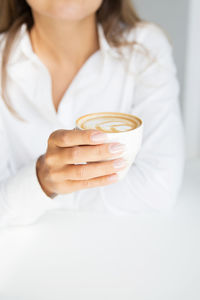 Woman in white shirt is holding cup of cappuccino coffee in day time, it is tender hand