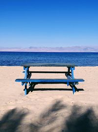 Empty bench on beach against clear blue sky