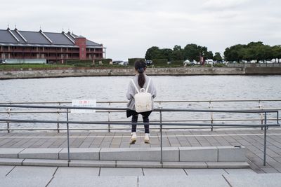 Rear view of woman sitting on railing by river against sky