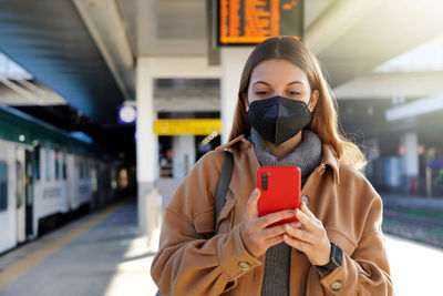 Stylish urban woman wearing black face mask kn95 ffp2 texting on smartphone in train station