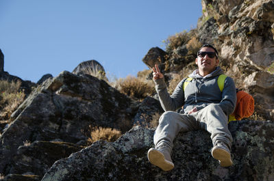 Low angle portrait of man gesturing while sitting on rock against mountains