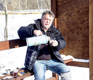 Man pouring tea while sitting on bench during winter
