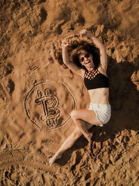 Portrait of young woman lying down by bitcoin symbol on sand at beach
