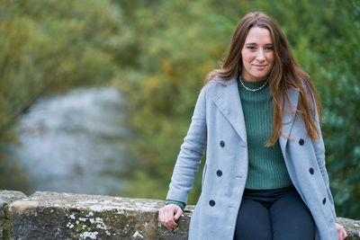 Portrait of a young caucasian woman with brown hair on a stone bridge with the green of the trees in the background. dressed in a gray jacket, green sweater, dark pants and military boots