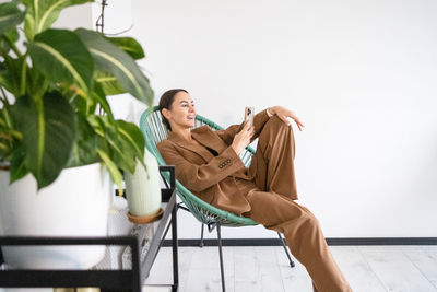 Young woman using phone and laughing at camera, she is wearing stylish suit and seating in chair