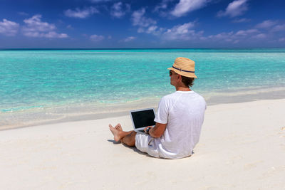 Rear view of man using laptop at beach against sky