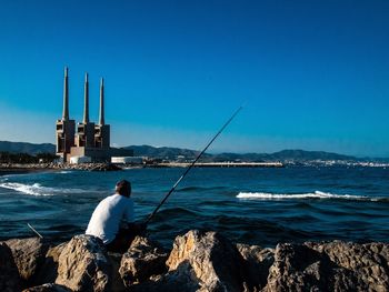 Rear view of man fishing in sea against clear blue sky