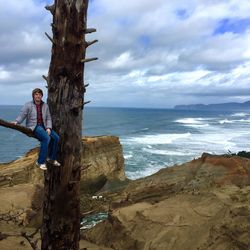 Young man sitting on tree branch at beach against cloudy sky