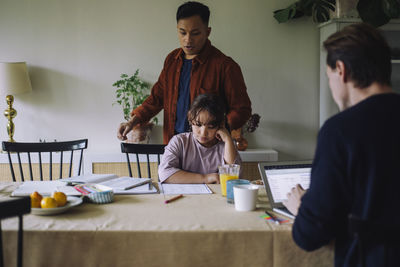 Gay couple parenting daughter studying while sitting at dining table in home