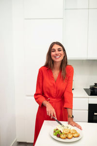 Charming young woman in red dress standing at kitchen counter and arranging plate with snacks while preparing food for home party