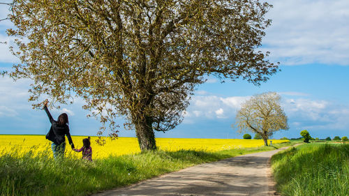 Rear view of mother and daughter standing with arms raised by tree against sky