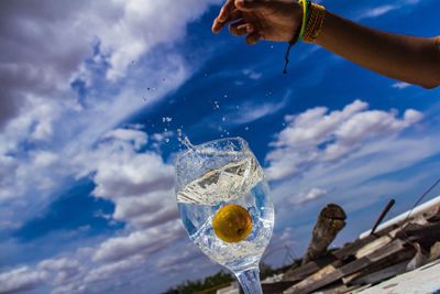 Cropped hand of person throwing lemon in drink against cloudy sky during sunny day