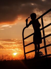 Silhouette boy standing on landscape against sky during sunset