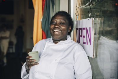 Cheerful female chef holding cup while standing by entrance sign