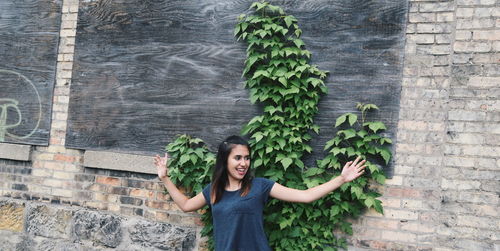 Cheerful woman standing with arms outstretched by plants growing on wall
