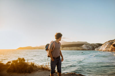 Anonymous male backpacker in casual clothes standing with hand in pocket and admiring scenic shore and rippling blue bay on sunny day in fyriplaka greece