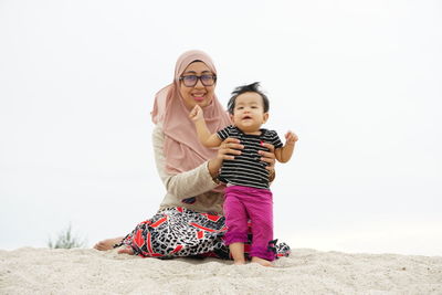 Mother wearing hijab holding cute daughter while sitting on sand at beach against sky