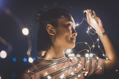 Close-up of smiling young woman with illuminated string lights in city at night