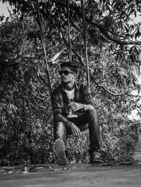 Full length of young man sitting on rope swing against trees