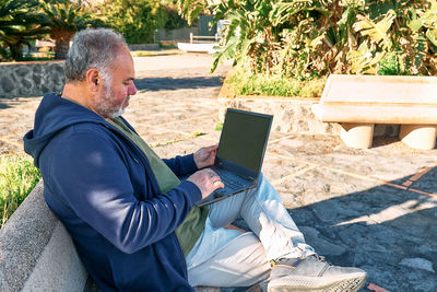 Handsome bearded mature man with laptop working outdoors while sitting on bench at the seaside. 