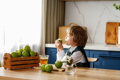 Thoughtful schoolboy is having breakfast with fresh juice and apple waiting for the school bus