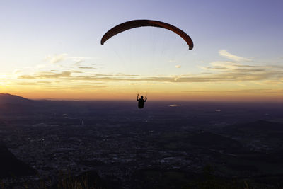 Silhouette person paragliding over landscape against sky at sunset