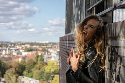 Portrait of young woman with blond hair showing rock sign against cityscape