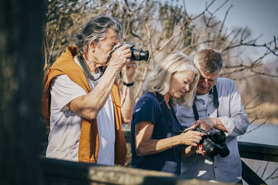 Senior friends using cameras while standing against bare trees at park