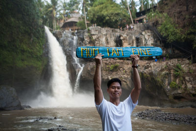 Portrait of man holding text on wood while standing against waterfall