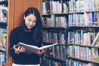 Young woman reading book while standing by bookshelves in library