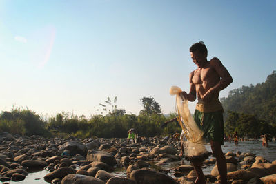 Full length of shirtless man standing on rock against sky