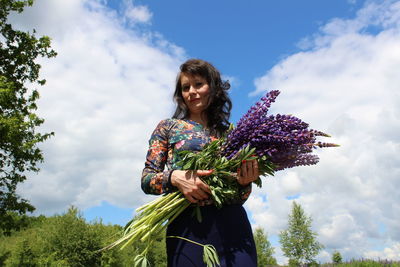 Low angle view of portrait woman holding lavenders while standing against cloudy sky