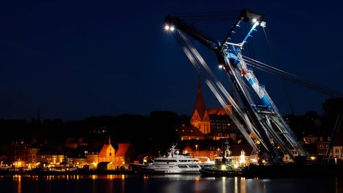 Illuminated shipping cranes at dock by town against sky