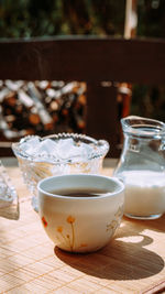 A cup of coffee stands outside on a table next to a jug of milk and a sugar bowl with lump sugar 