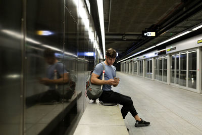 Young man using smart phone while sitting on bench against wall at illuminated subway