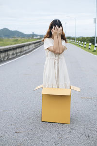 Full length of woman covering face standing in cardboard box on road