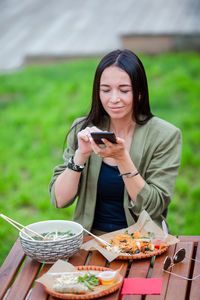 Beautiful smiling woman using phone eating food while sitting outdoors