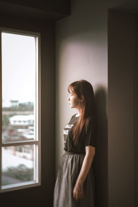 Side view of young woman looking through window at home