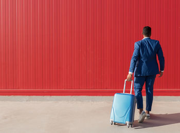 Full body back view of unrecognizable ethnic businessman in elegant formal suit pulling suitcase while walking against red wall