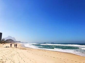 People against clear blue sky at beach on sunny day