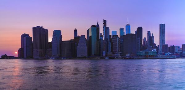 Sea by modern buildings against sky during sunset